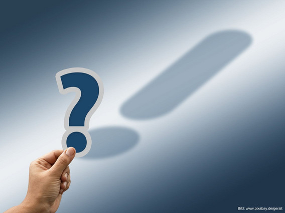 Hand hält ein Fragezeichen, welches ein Ausrufezeichen als Schatten wirft.